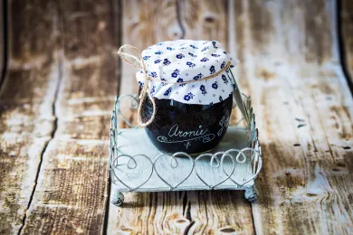 Tmavá skleněná lahev bylinného sirupu s látkovým víkem modrý květinový vzor a jutový provázek na kovovém stojánku na dřevěném stole